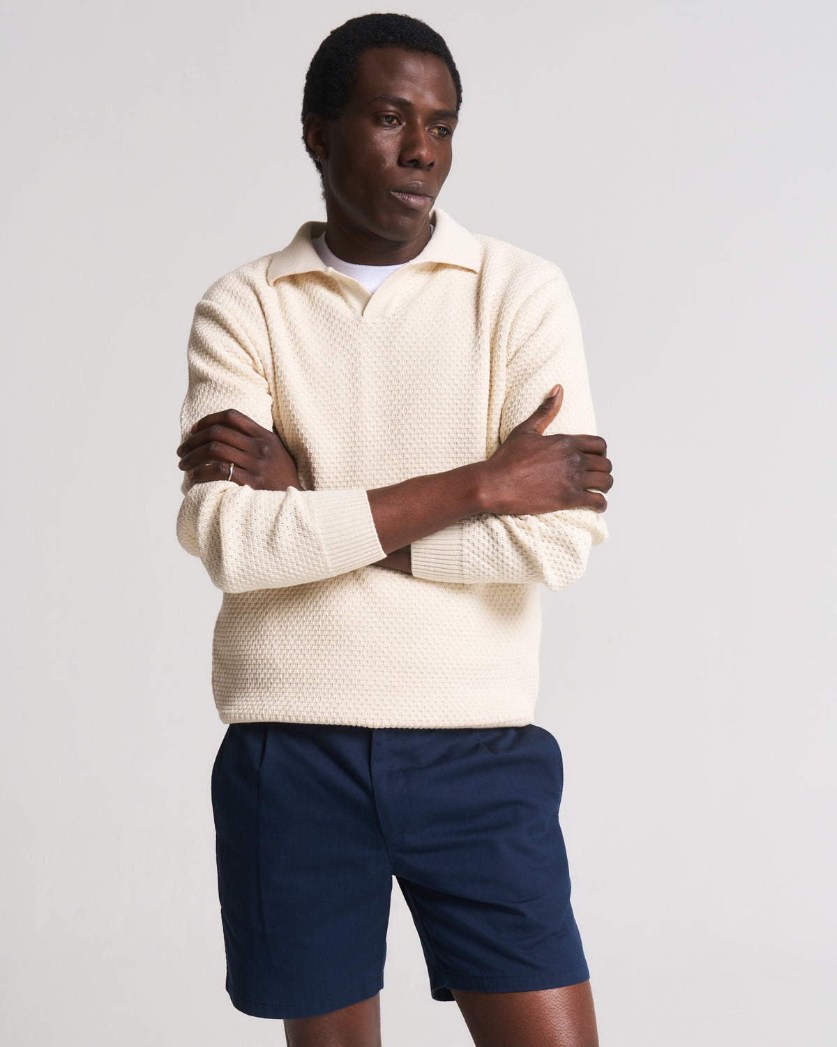 A man wearing a cream-colored, textured knit polo sweater and navy blue shorts, standing with arms crossed against a light gray studio background.