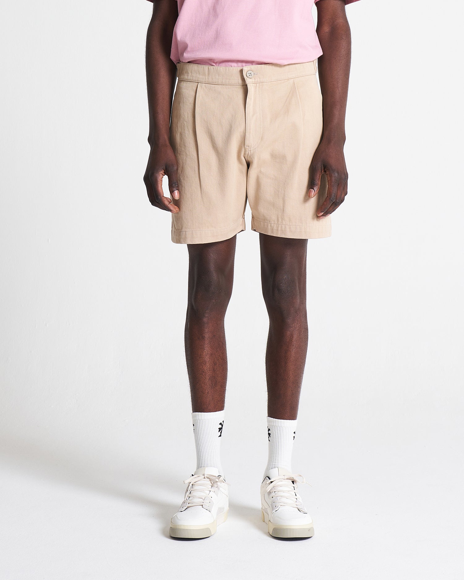 A person standing wearing beige relaxed cotton shorts and a pink top, paired with white socks and white sneakers.