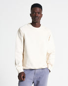 A man wearing an off-white crew neck sweater with rib-knit finishes at the neckline, cuffs, and hem. The sweater is made from cotton and has a soft appearance.