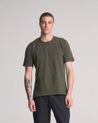 Man wearing an olive green knitted t-shirt and black pants on a light gray background