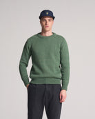 Man wearing a olive green sweater and dark pants against a plain background