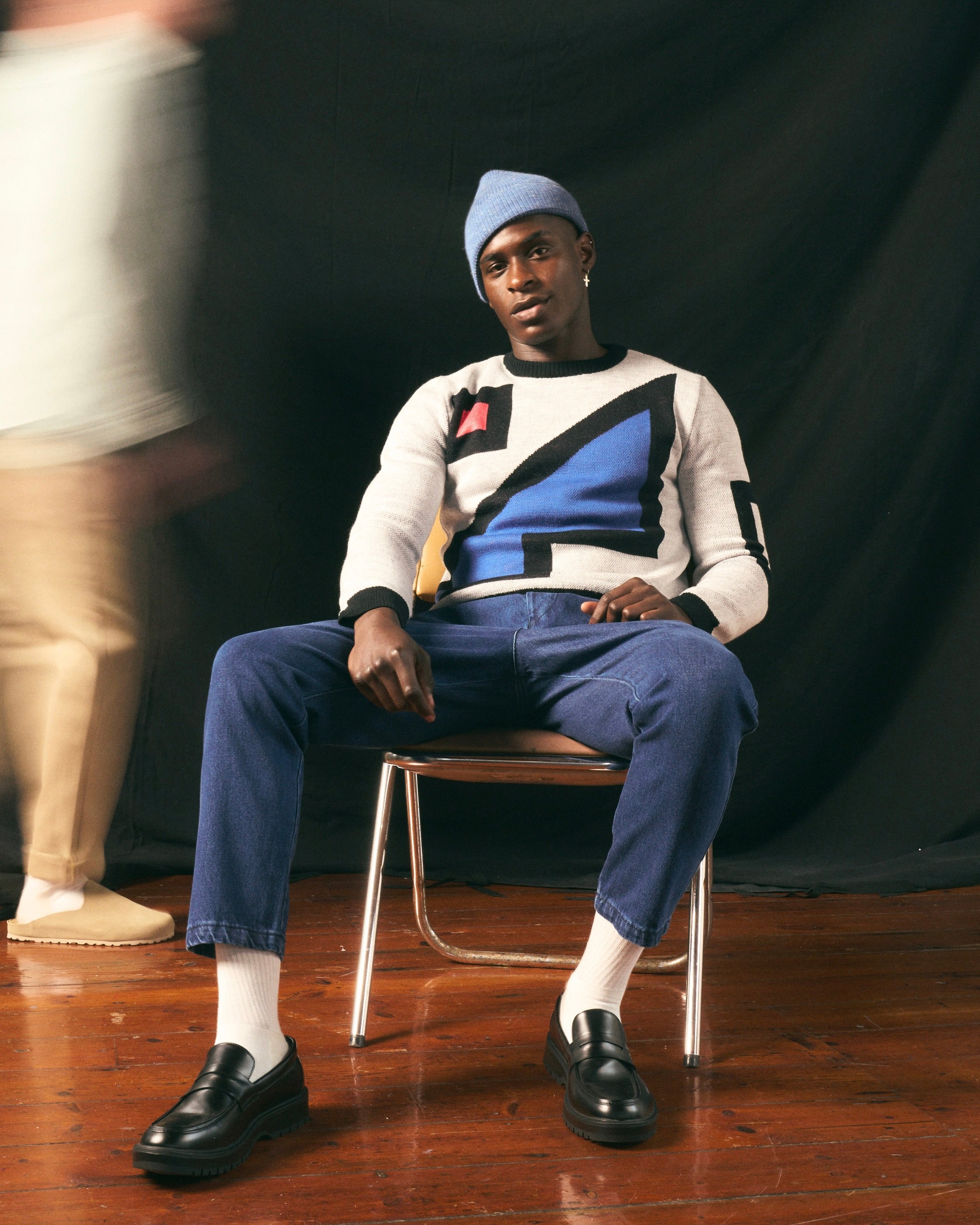 Person sitting on a chair wearing a patterned sweater featuring the artwork of The Ninevites and blue pants against a black background.