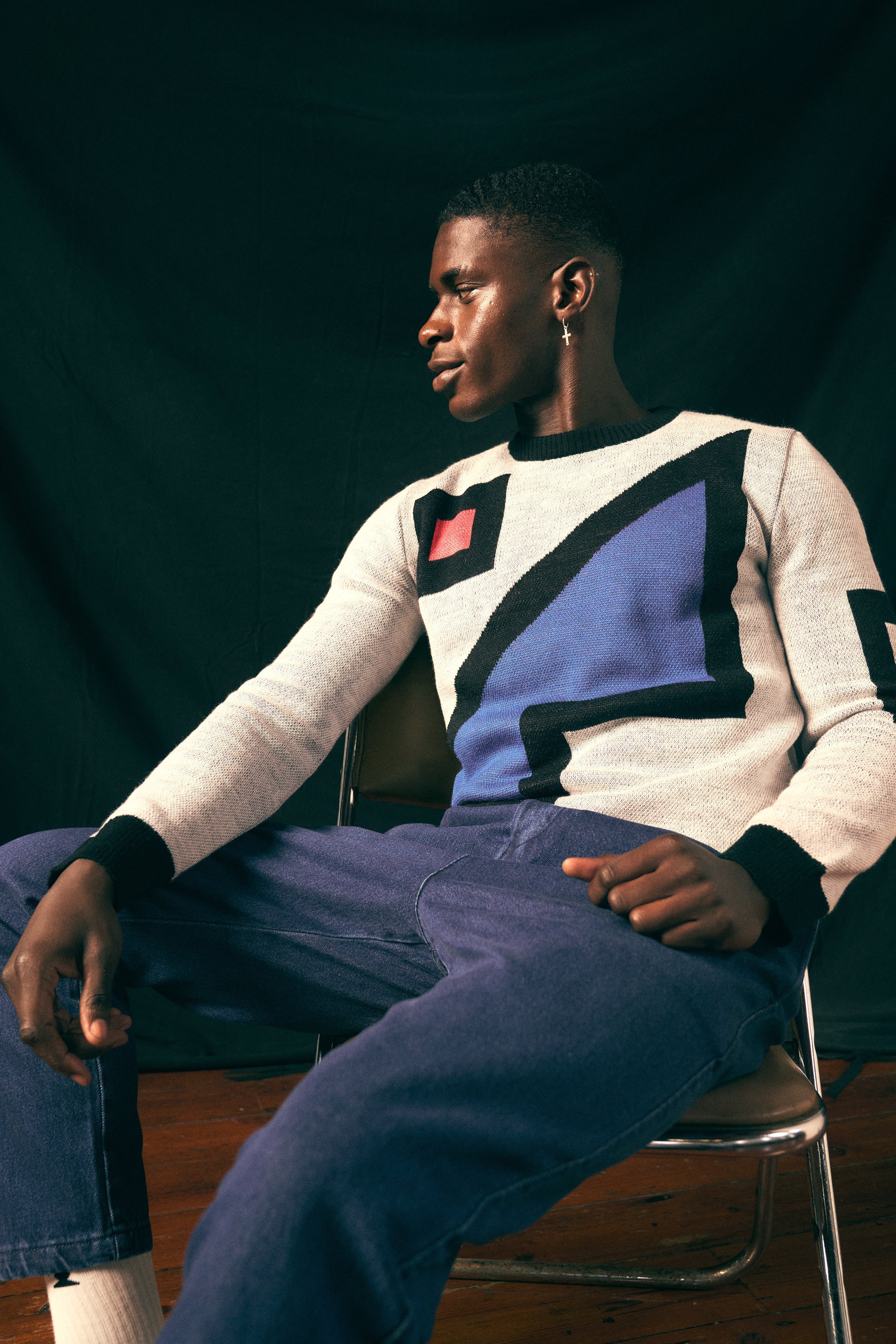 Person wearing an artist collaboration sweater sitting on a chair against a dark background. By Fields.
