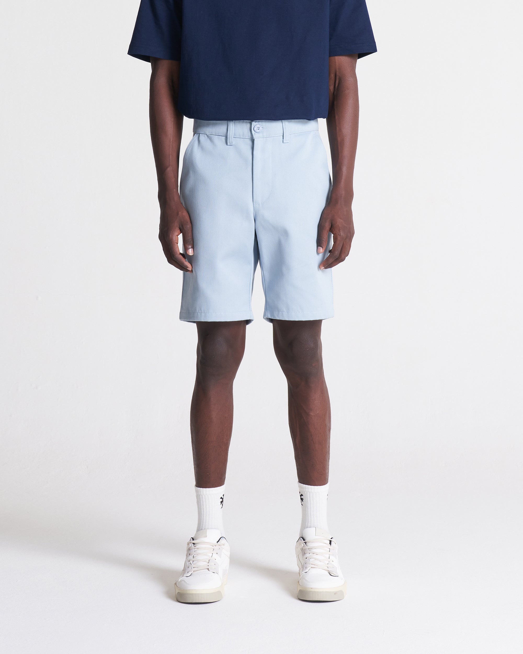 A person standing wearing light blue cotton twill art shorts with a fitted silhouette, including front quarter-slant pockets, a single coin pocket, back welt pockets, and a zip fly.
