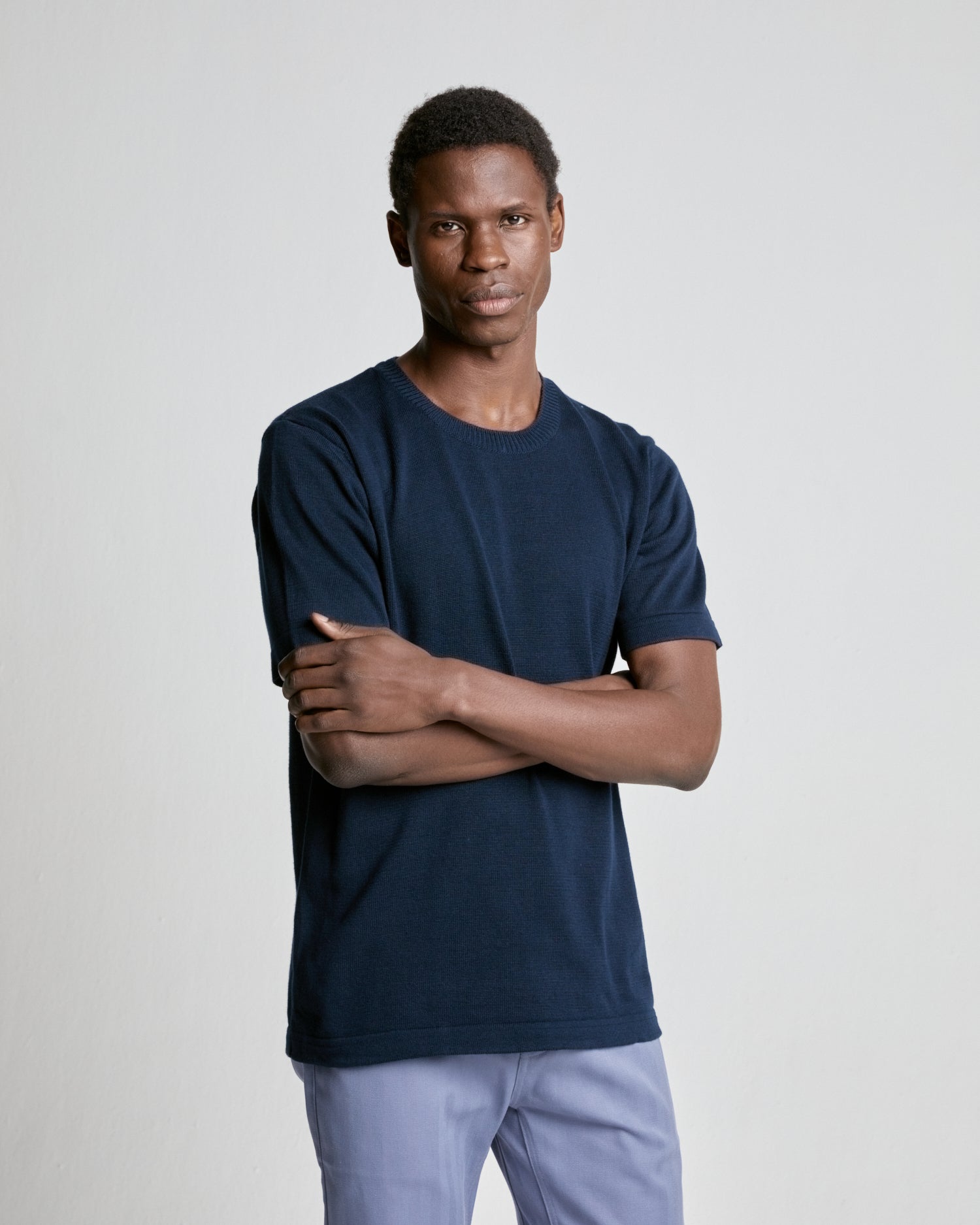 A person wearing a navy knitted tee and light trousers, standing with arms crossed.