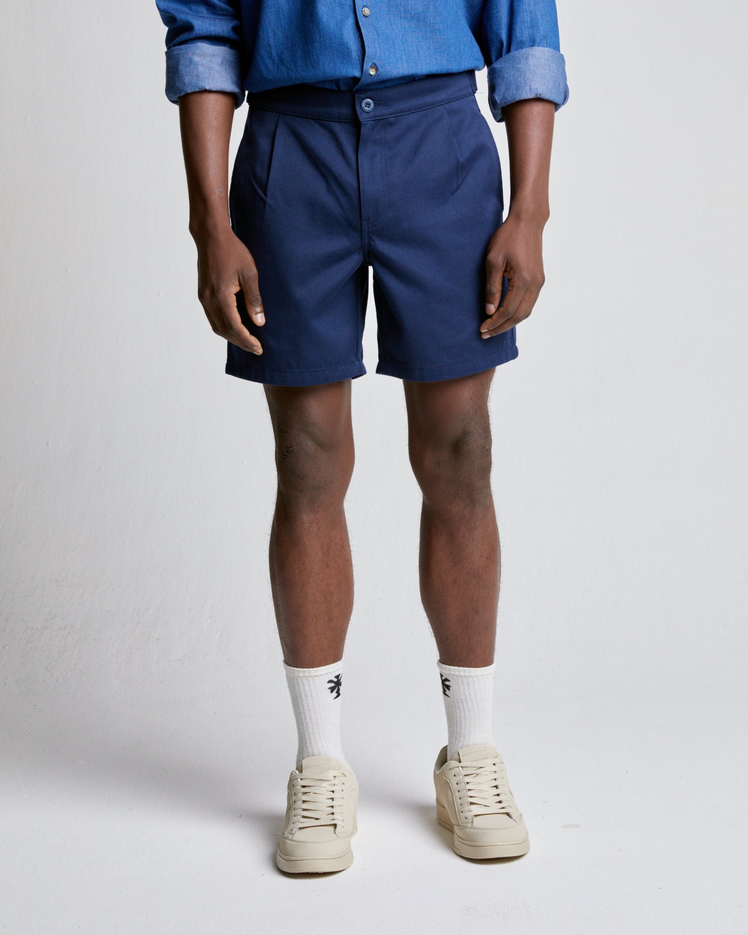 A person standing wearing relaxed fit navy cotton shorts, a blue shirt, white socks, and beige shoes.