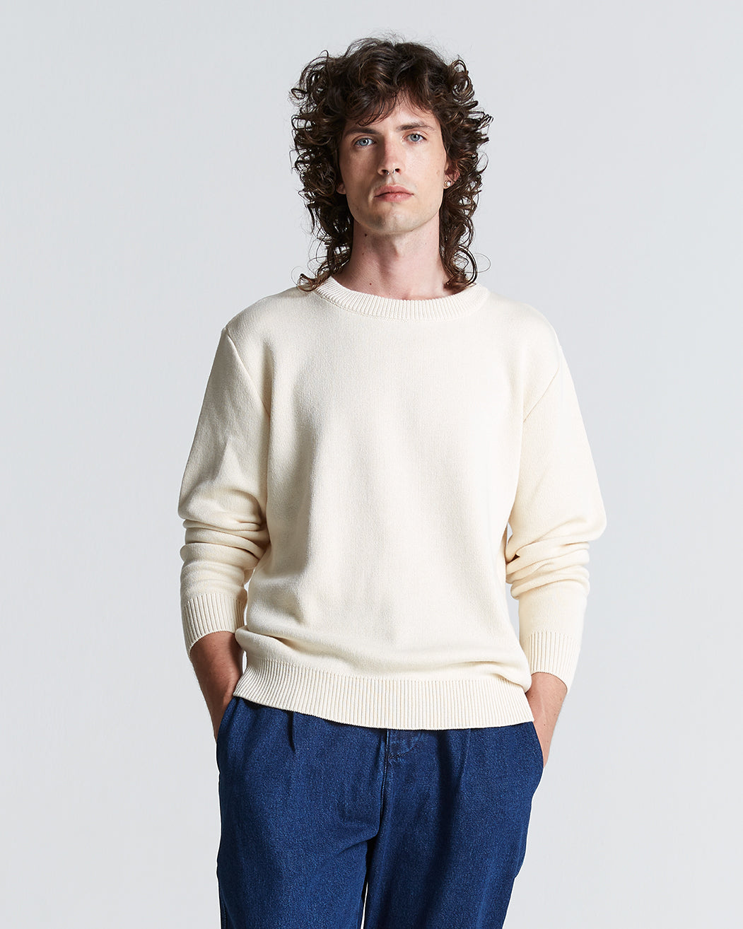 A person with curly hair wearing a round neck, cream-colored sweater in cotton and blue pants.