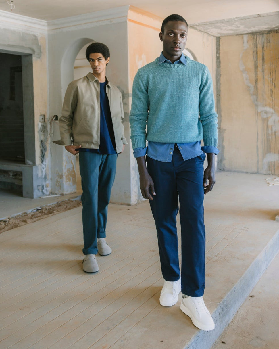 Two male models walking down a set of concrete steps in an unfinished building with columns. The model in the foreground is wearing a light blue knit sweater over a blue collared shirt, navy trousers, and white sneakers.