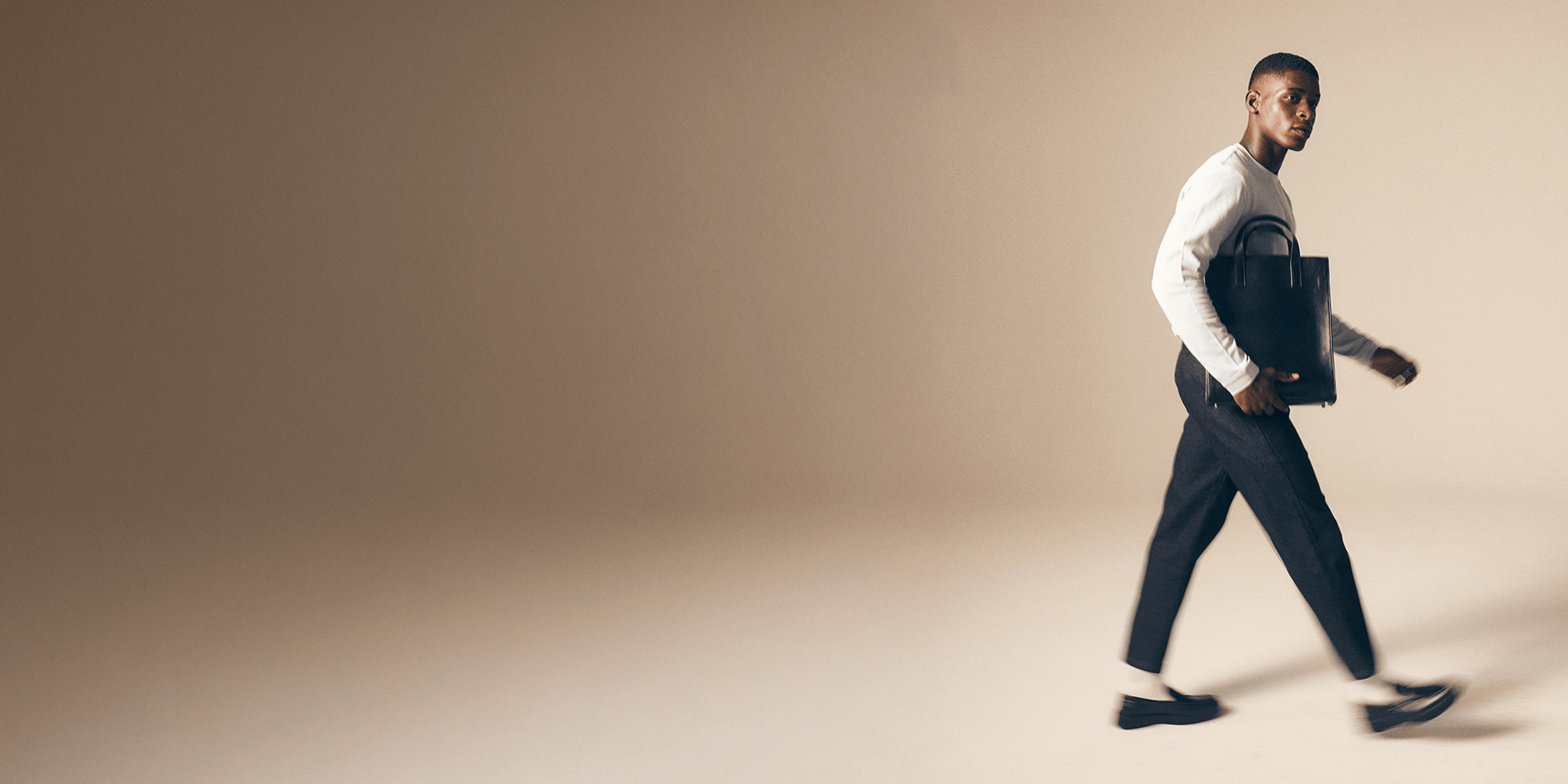 Man walking wearing Fields pique t-shirt, denim trouser, with a briefcase against a beige background