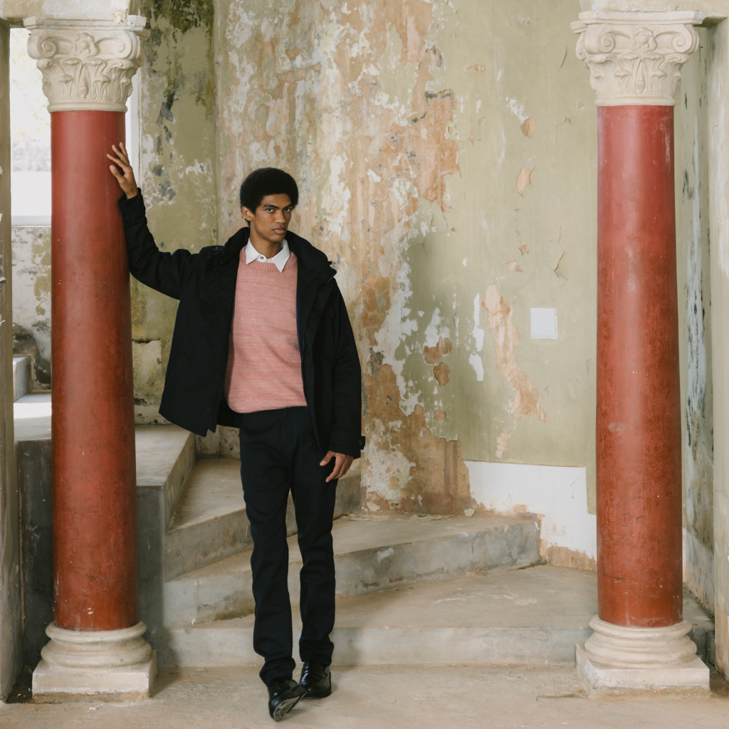 A man wearing a black jacket, pink sweater vest, white collared shirt, and black trousers stands on stone steps between two red columns in a rustic, peeling-wall interior.