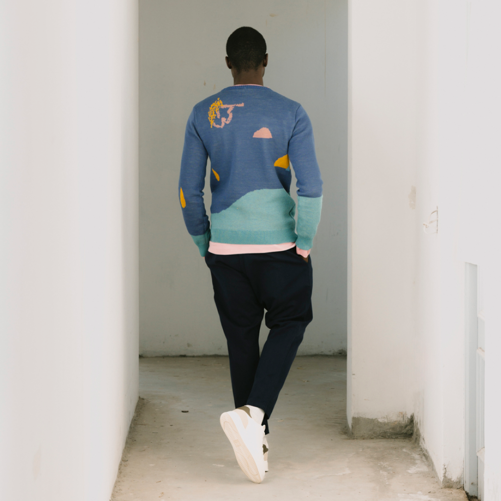 A man viewed from behind walking down a white hallway, wearing a color-blocked knit sweater with blue, teal, and yellow sections, dark pants, and white sneakers.