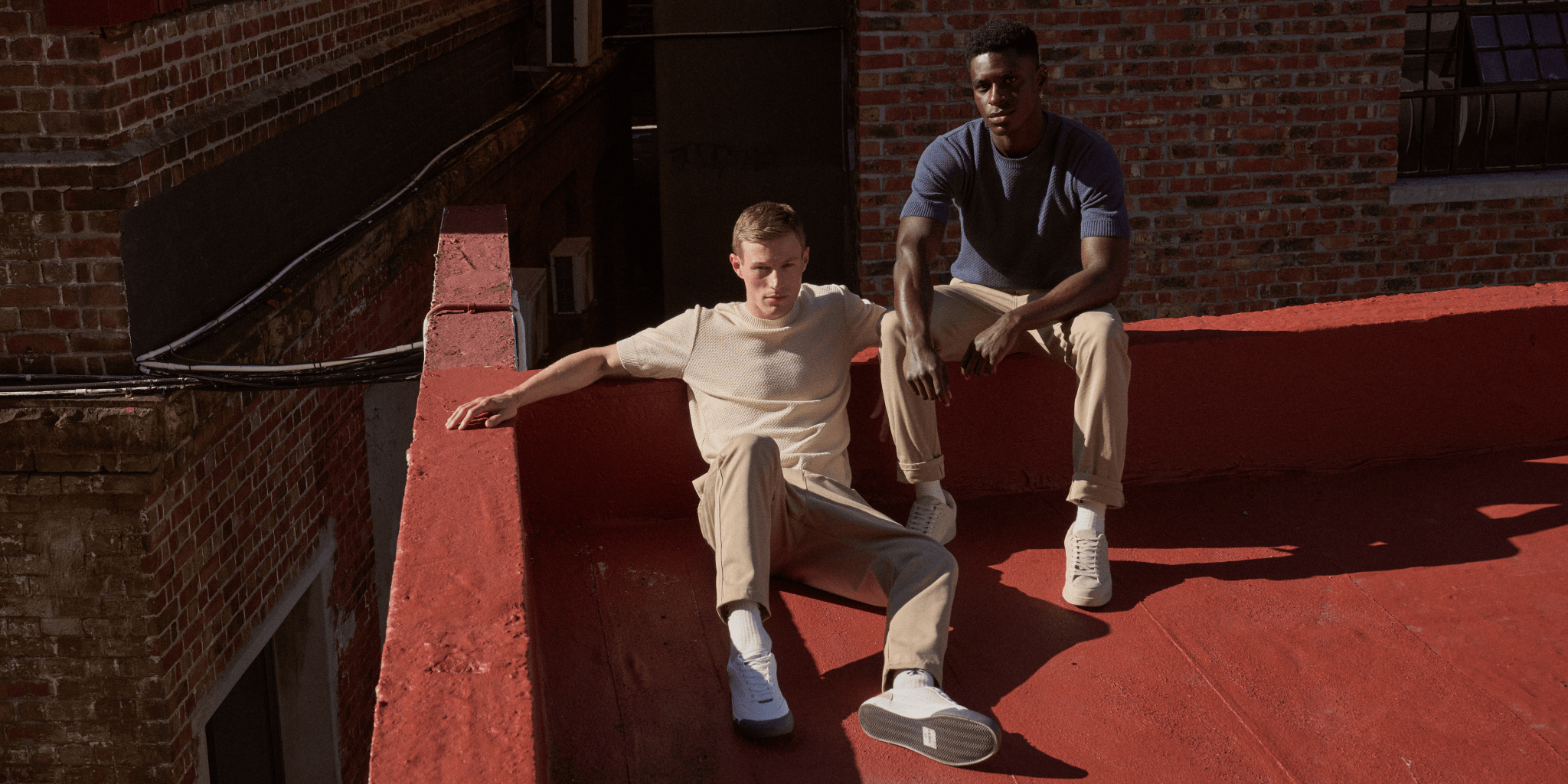 Two men sitting on a red ledge against a brick wall wearing Fields clothing