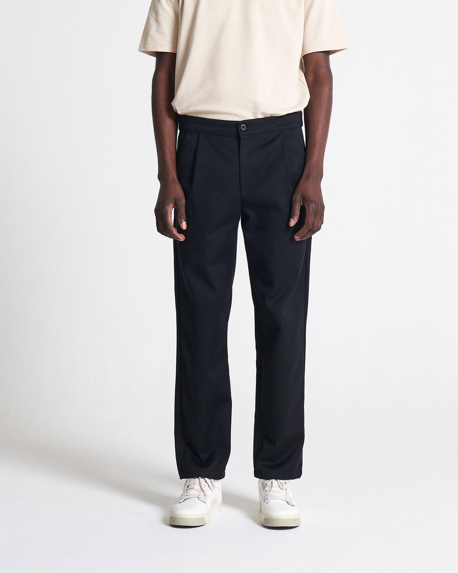 A person standing wearing black straight-leg trousers and a beige cotton t-shirt.