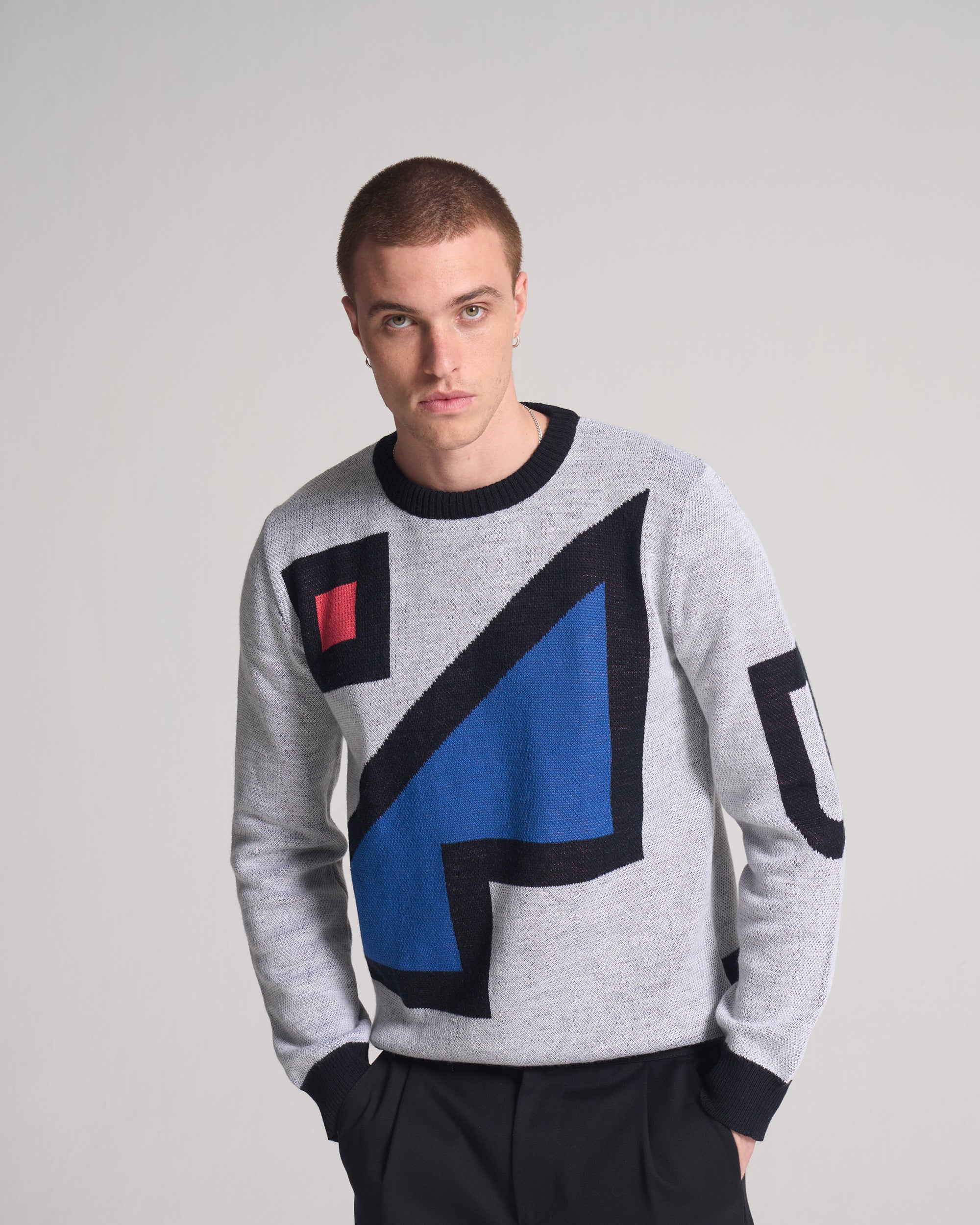 Man wearing a grey sweater with geometric colour blocks on a plain background. Sweater is an artist collaboration limited edition sweater. 