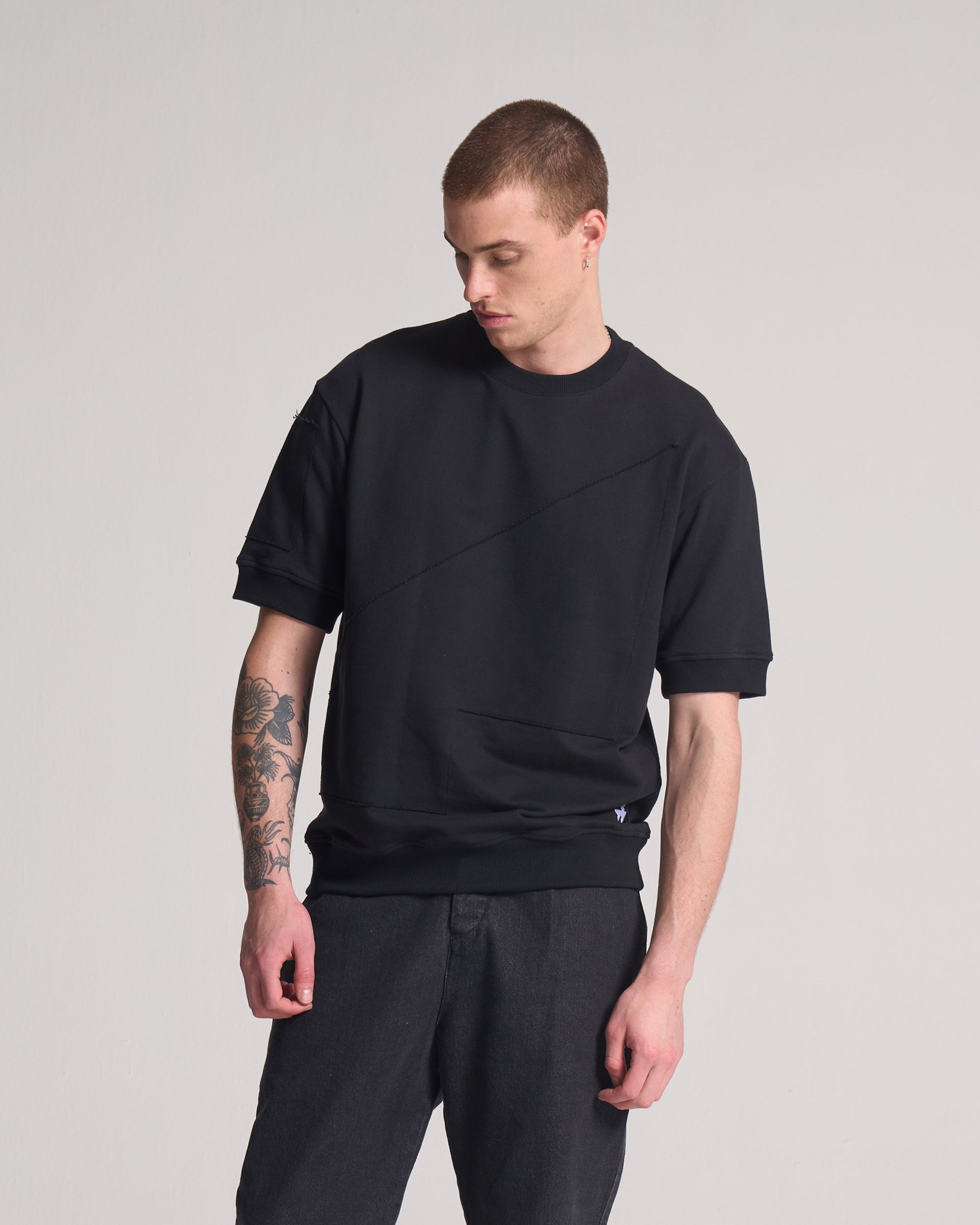 Man wearing a black short sleeve sweater and pants on a gray background. Sweater shows detail for an artist collaboration.