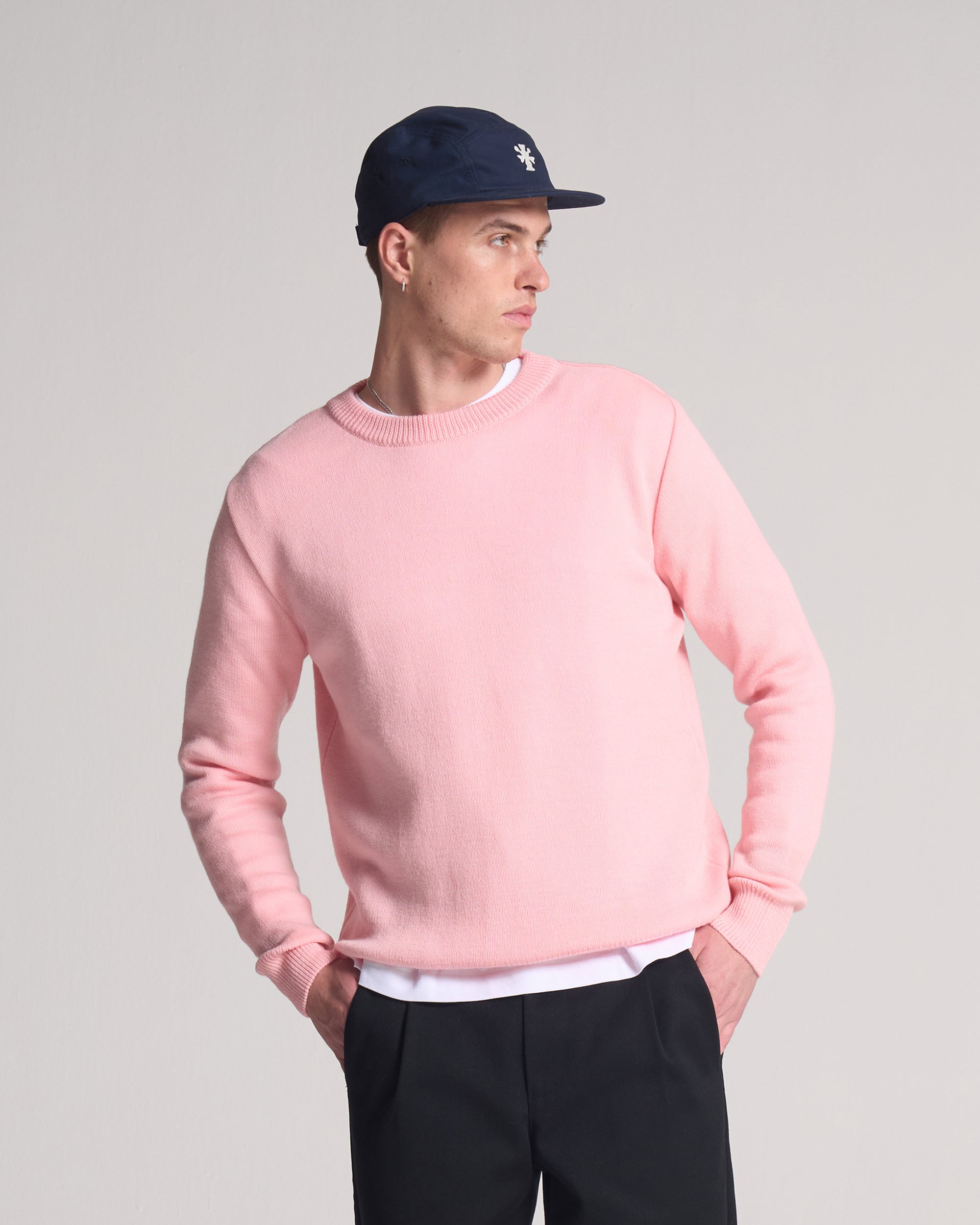 Person wearing a pink sweater made in wool and mohair, navy cap, and black pants on a grey background