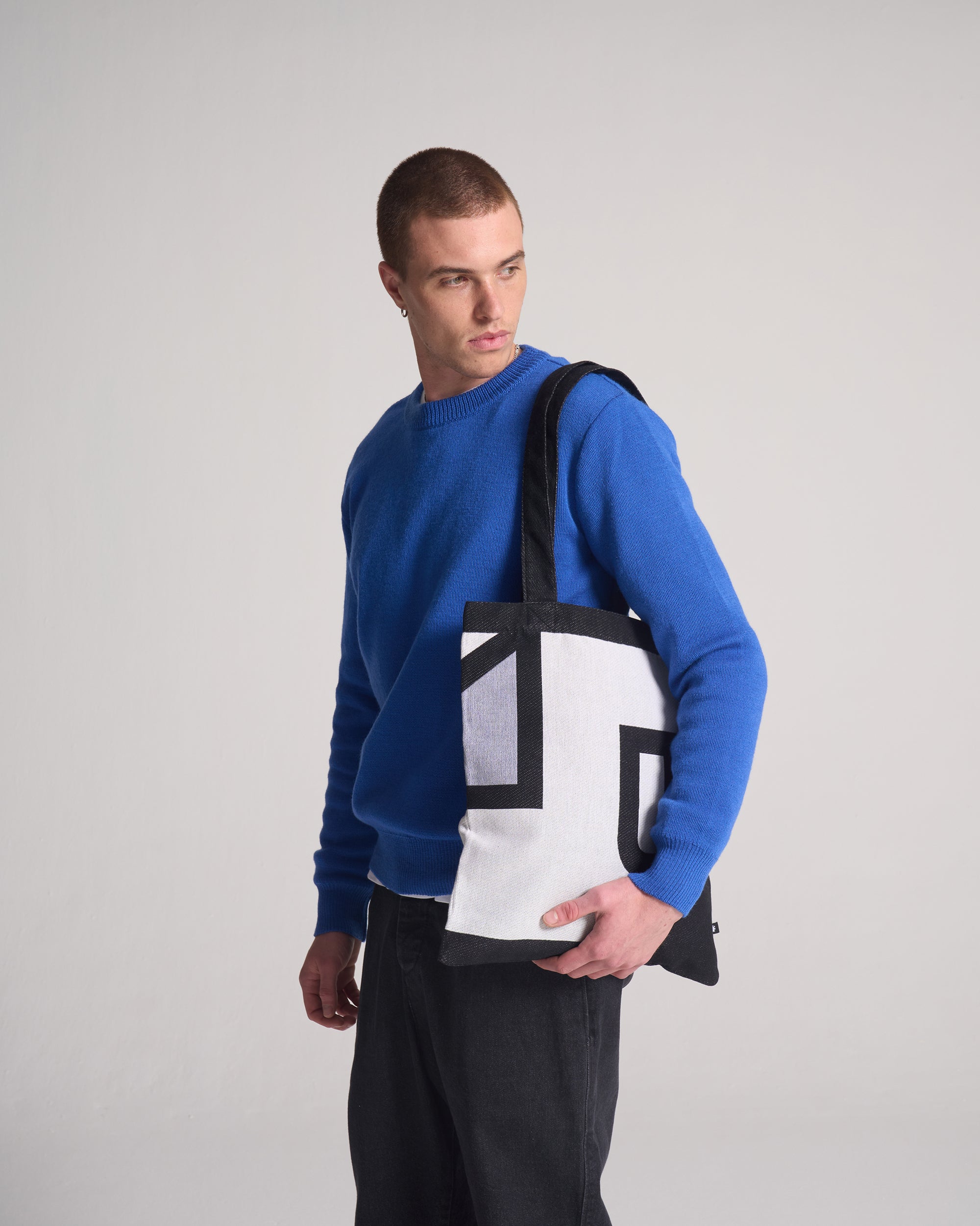 Man wearing a blue sweater holding a an artist collaboration black and white bag against a gray background