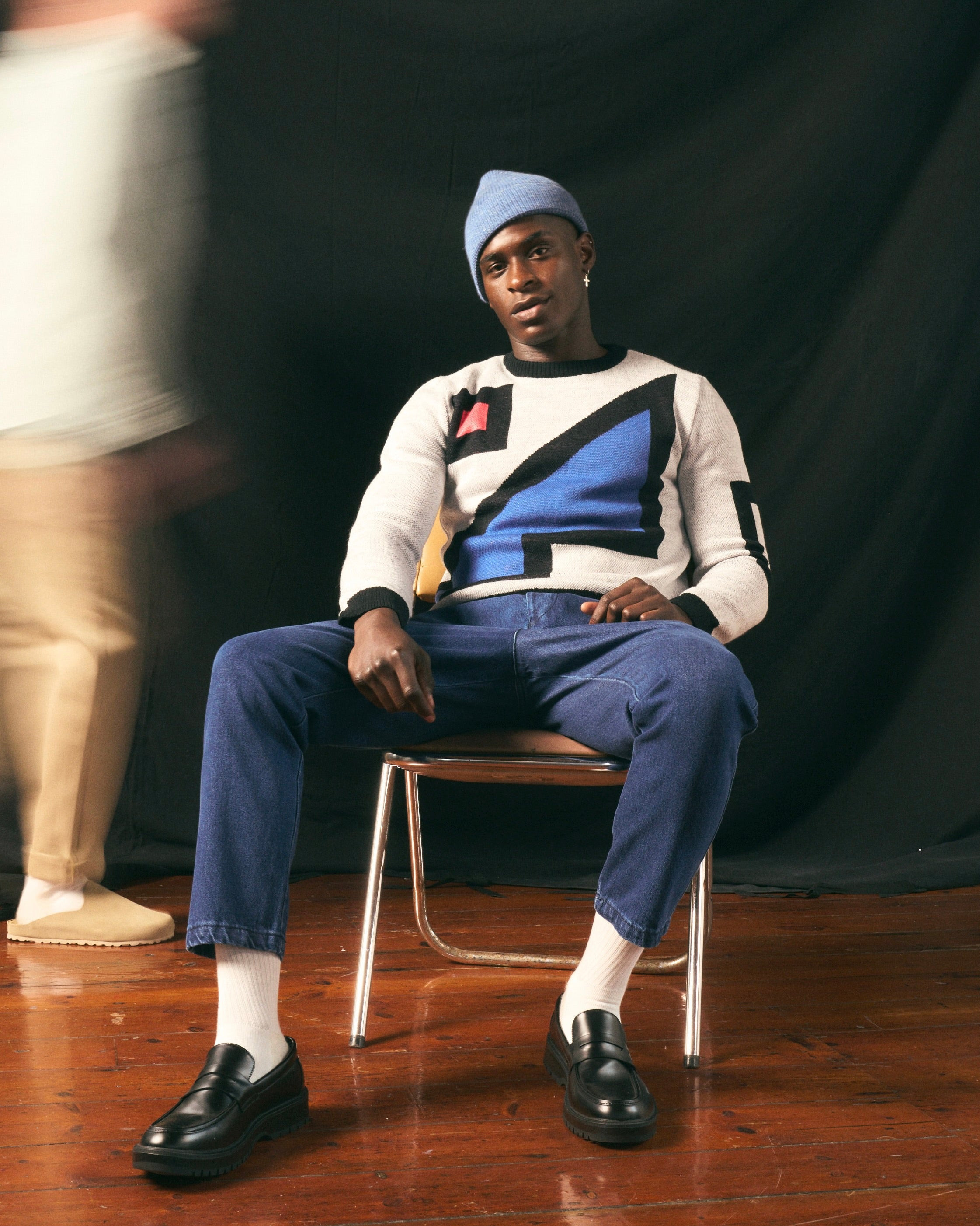 Person sitting on a chair wearing a patterned sweater featuring the artwork of The Ninevites and blue pants against a black background.