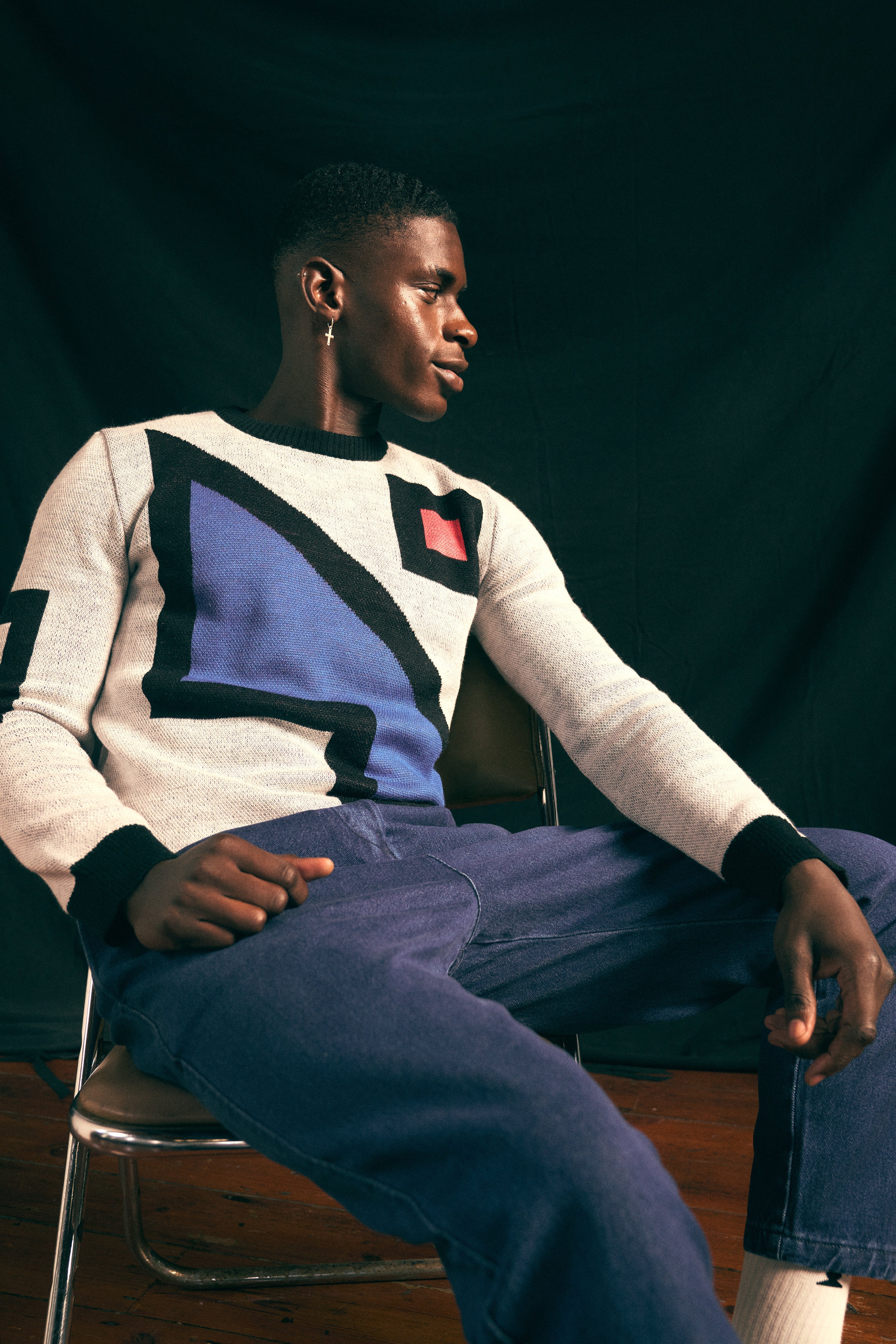 Person wearing an artist collaboration sweater sitting on a chair against a dark background. By Fields.