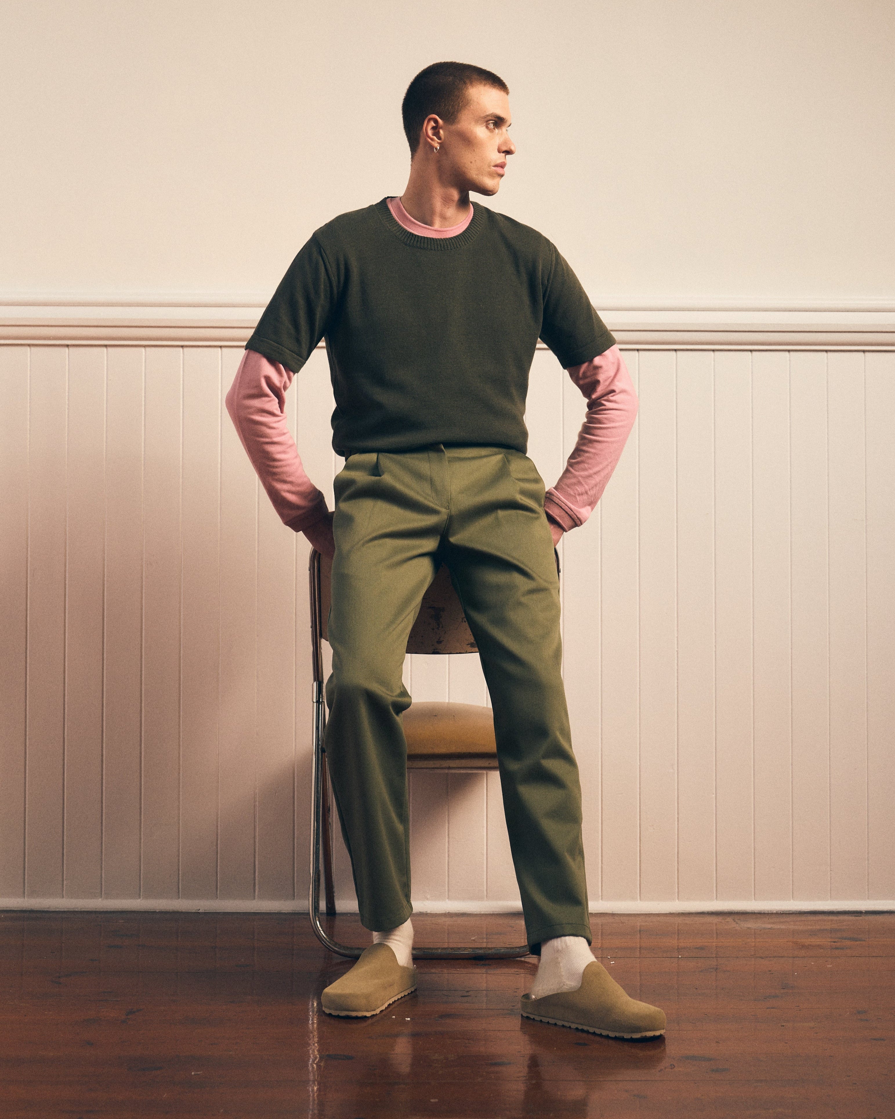 Man wearing a green knitted tee, over a pink pique tee and khaki pants standing against a plain wall.