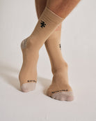 Beige socks with black logo worn by a person on a white background. Crew height.