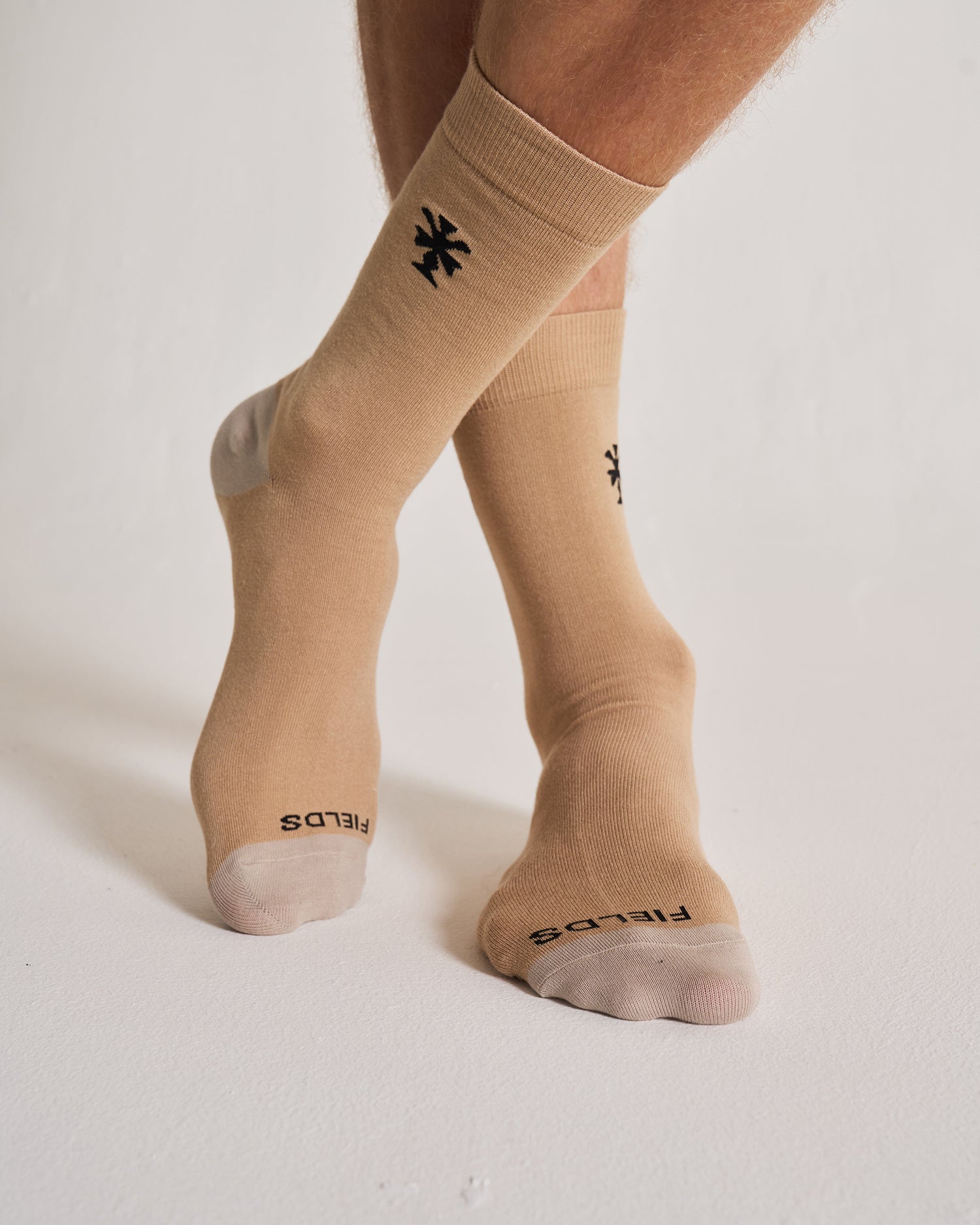Beige socks with black logo worn by a person on a white background. Crew height.