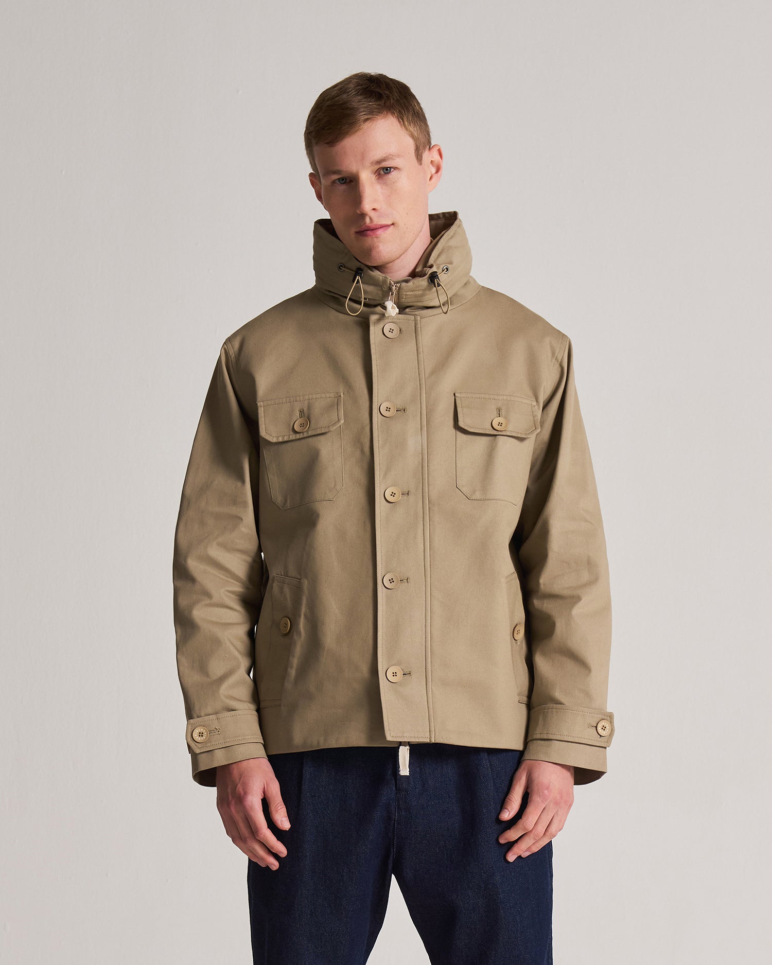 Man wearing a beige Field jacket on a plain background. Jacket features multiple pockets and a secret hood in the neckline. 