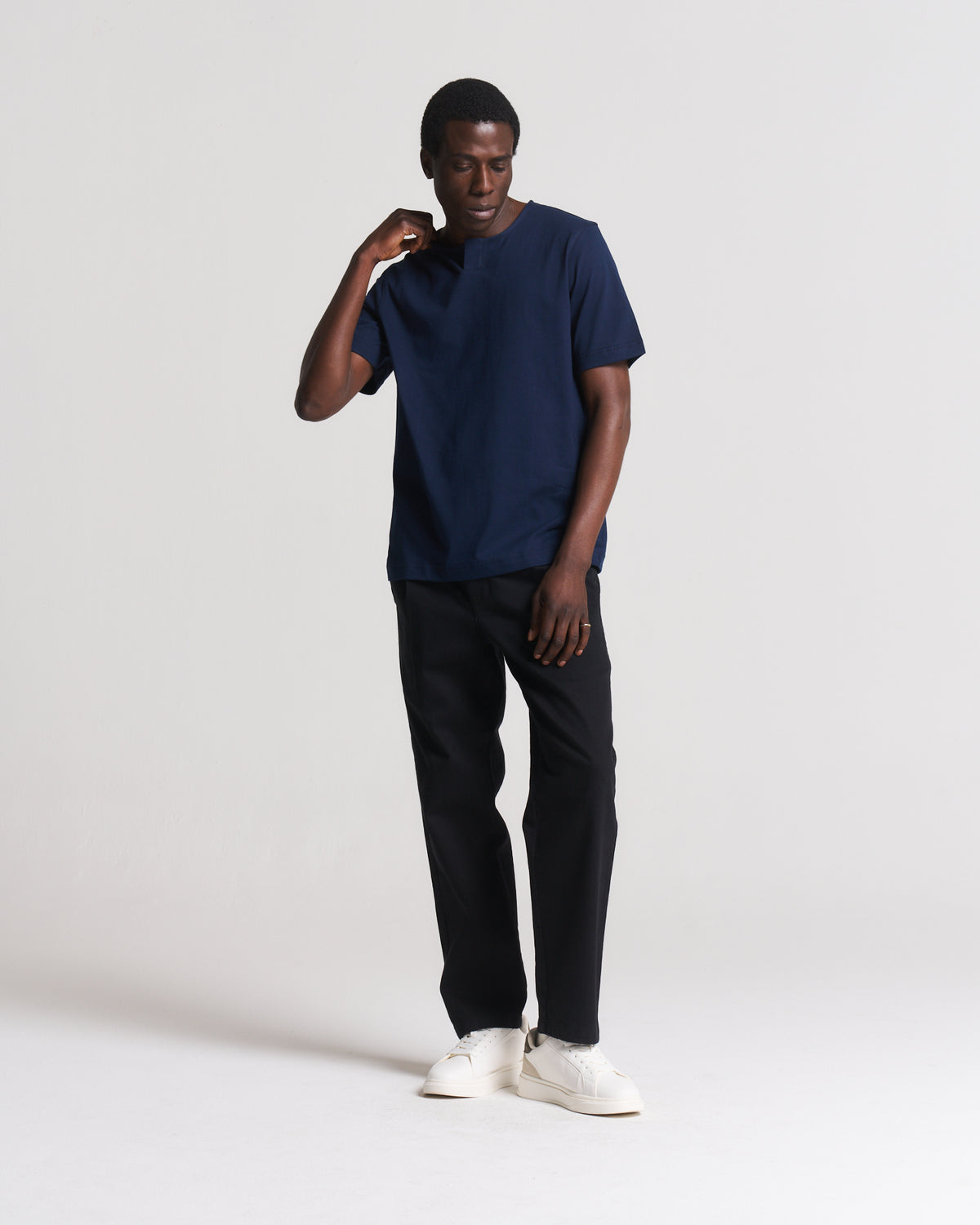 A man modeling a navy blue short-sleeve crewneck t-shirt, paired with black trousers and white sneakers, standing against a light gray studio background.