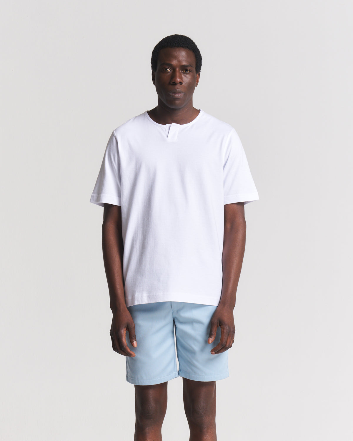 A man modeling a white short-sleeve t-shirt with a split crew neck and light blue shorts, standing against a light gray studio background.