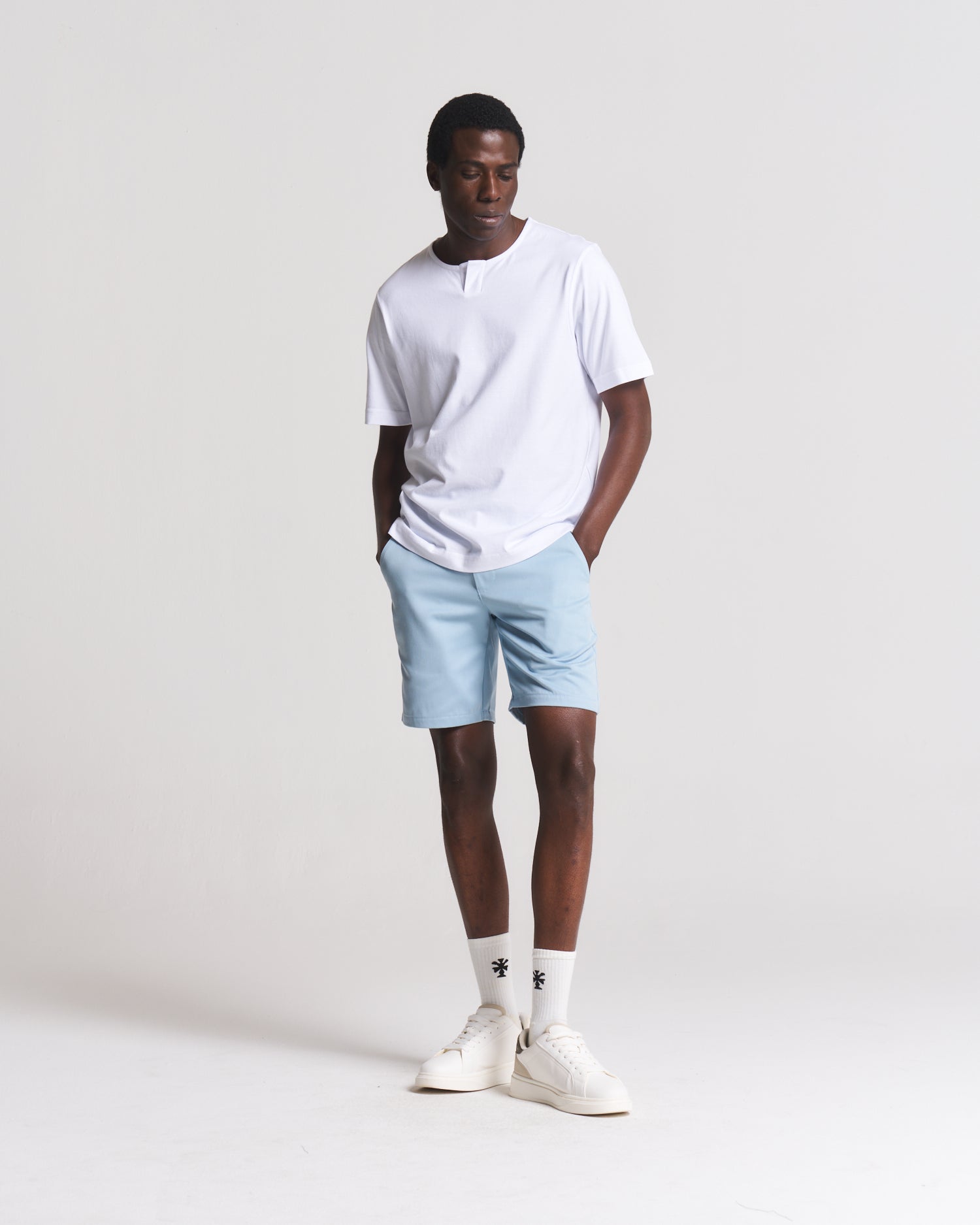 A man modeling a white short-sleeve henley t-shirt, light blue shorts, white crew socks, and white sneakers against a plain light gray studio background.