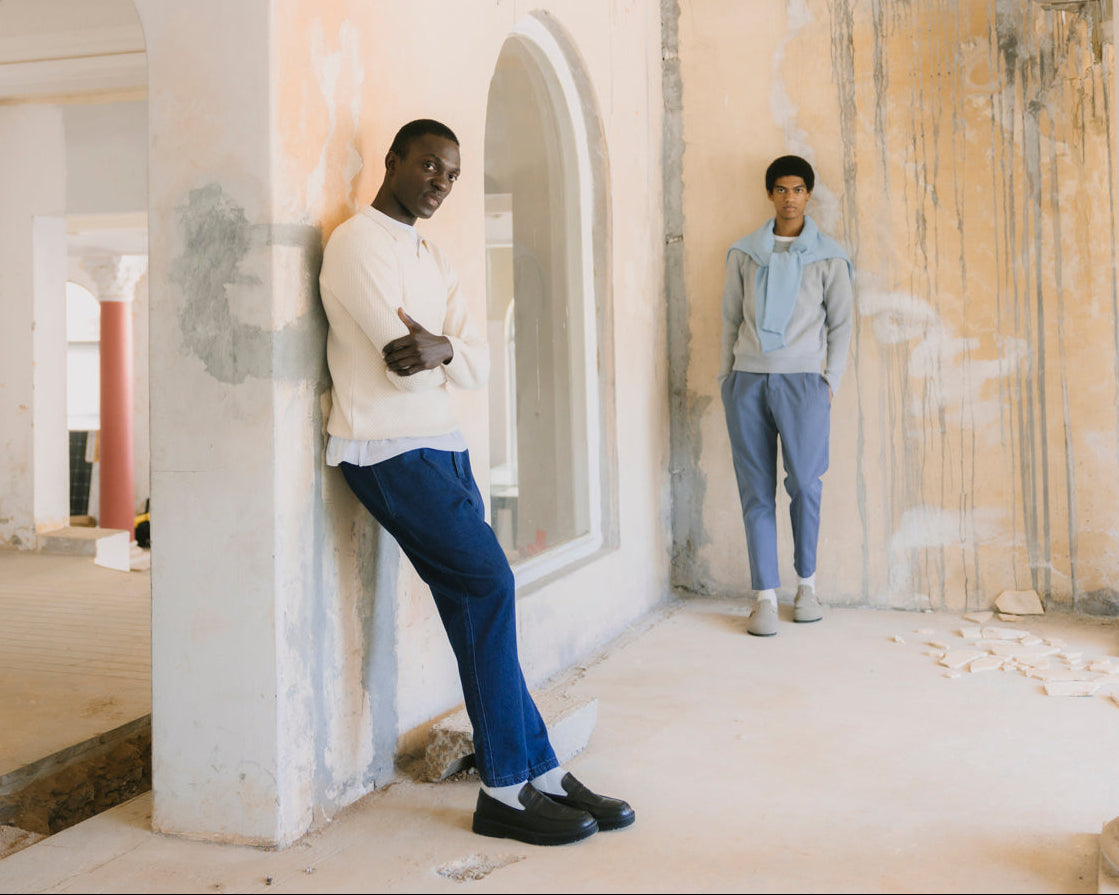 Two models posing in an unfinished building with plaster arches and walls. The model on the left wears a cream long-sleeve shirt and blue trousers, leaning against a pillar.