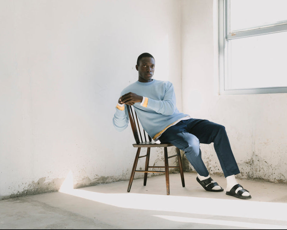 A man sits on a wooden chair in a bright, minimalist room with white walls and a large window. He is wearing a light blue long-sleeve knit sweater with yellow and white striped cuffs, dark blue jeans, and black slide sandals.