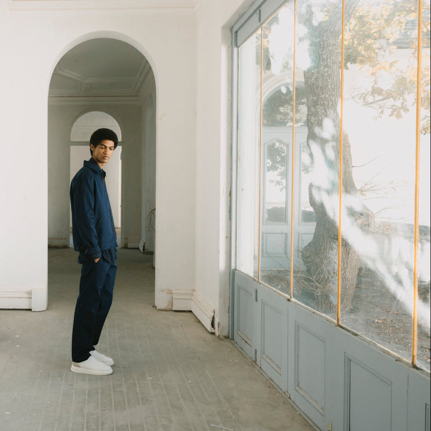 A man wearing a dark navy blue jacket and white sneakers stands in a bright, white corridor with arched doorways and large windows.