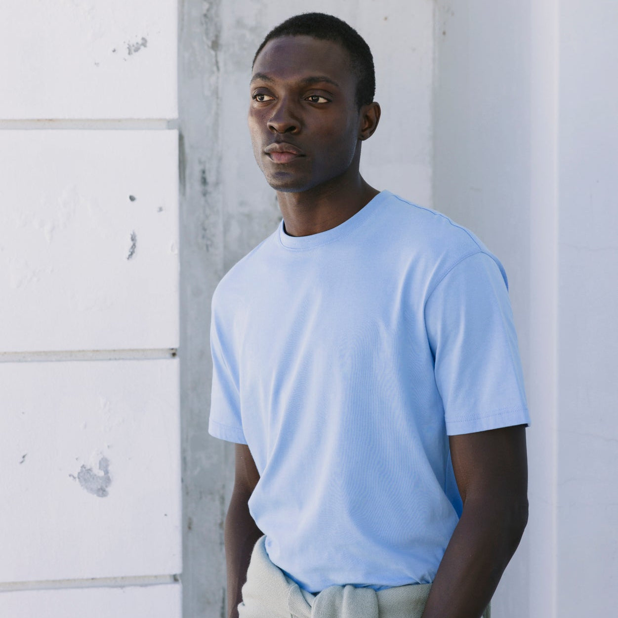 A man wearing a light blue crewneck t-shirt and blue pants, standing against a white textured wall.