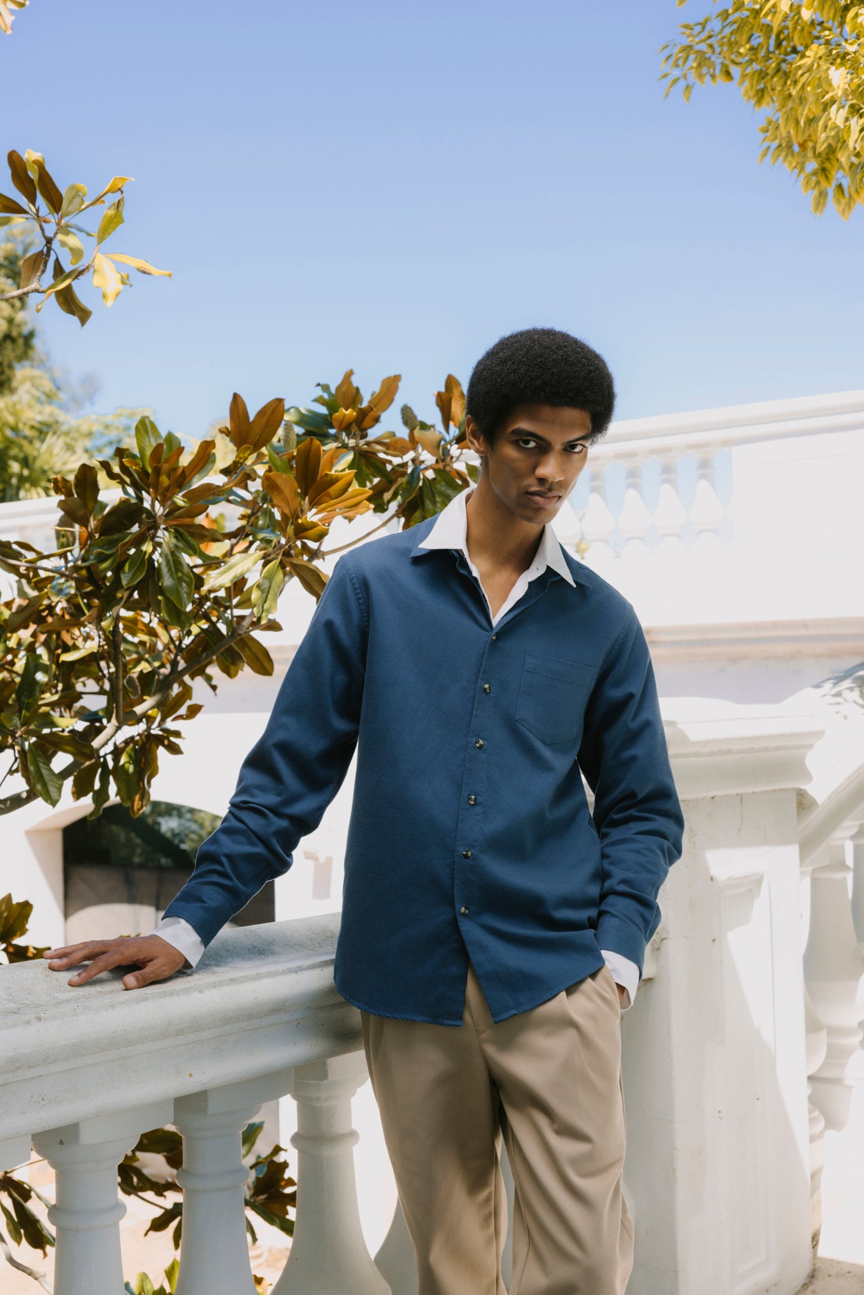 A man wearing a navy blue long-sleeve button-down shirt and beige trousers, leaning against a white outdoor railing under a clear blue sky.