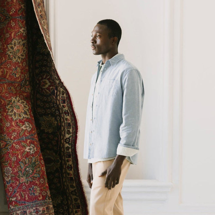A man in a light blue button-down shirt and beige pants stands in a bright white room, looking at a large rolled red patterned rug leaning against the wall.