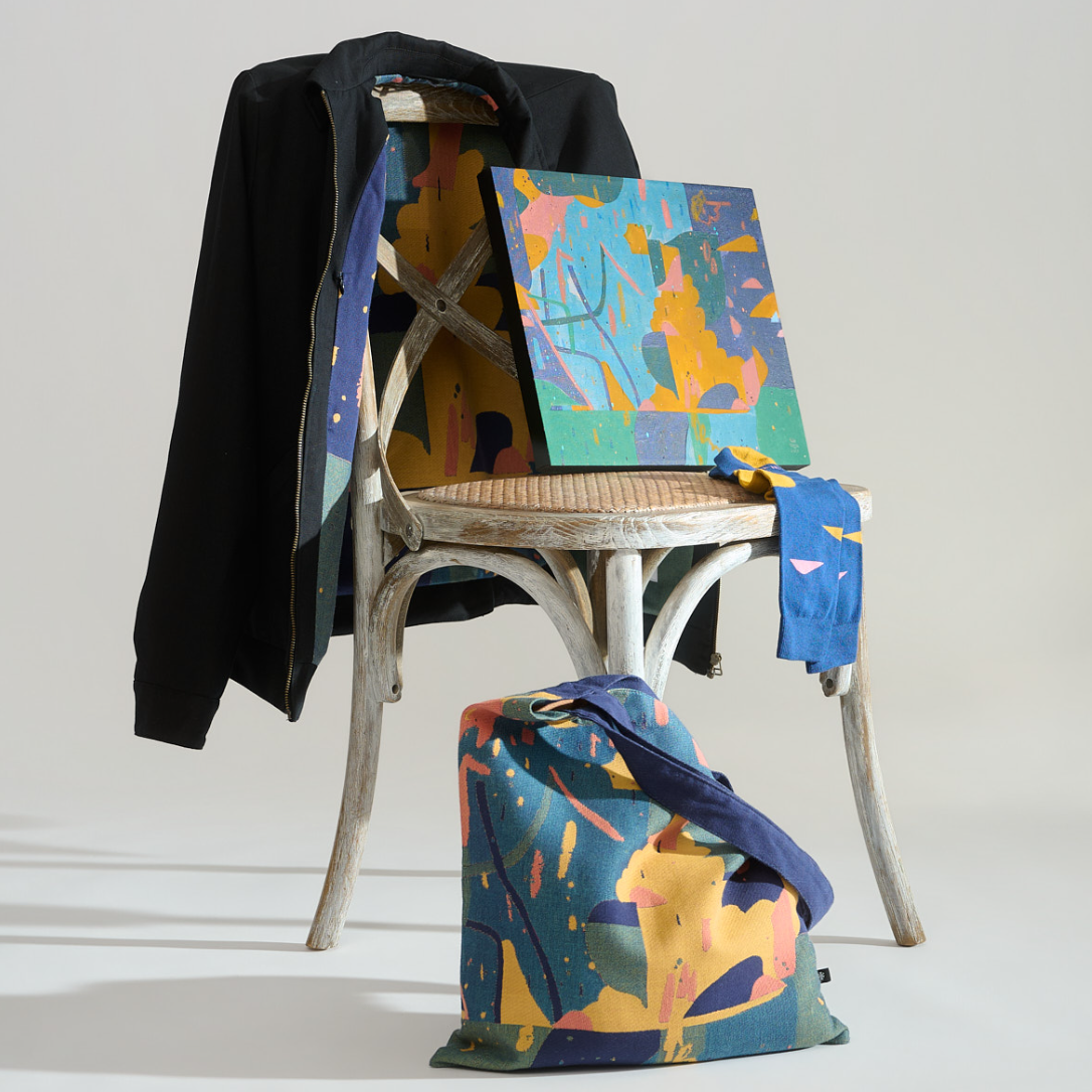 A styled studio shot featuring a matching multicolor abstract print set across a black cardigan draped over a rustic wooden chair, a matching square canvas print leaning on the chair, a matching blue scarf draped over the arm, and a matching tote bag on the floor. The print combines shades of blue, yellow, teal, and orange. WIth FIELDS x Paul Senyol pieces.